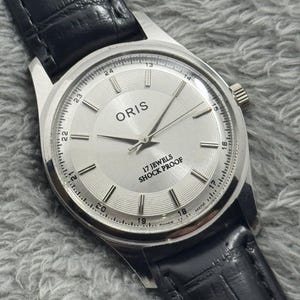 以下が含まれることがあります： 黒い革バンドが付いた、シルバーカラーのORIS腕時計。文字盤はシルバーで、黒い針と時間マーカーが付いています。「ORIS」と「17 JEWELS SHOCK PROOF」の文字が見えます。バンドは黒色です。