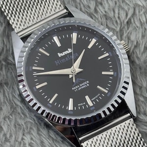 Puede incluir: Reloj de pulsera plateado con esfera negra. El reloj muestra el texto "hmt Himalaya" y "Para Shock 17 Jewels". Tiene una correa de malla y un bisel plateado.