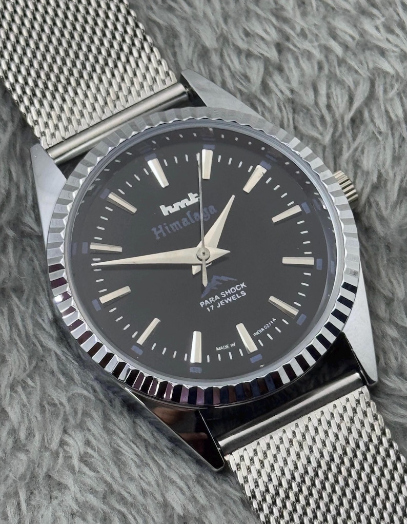 Puede incluir: Reloj de pulsera plateado con esfera negra. El reloj muestra el texto "hmt Himalaya" y "Para Shock 17 Jewels". Tiene una correa de malla y un bisel plateado.