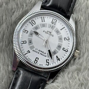 K&ouml;nnte beinhalten: Eine silberfarbene Fortis-Uhr mit wei&szlig;em Zifferblatt, schwarzen Zeigern und einem schwarzen Lederarmband. Das Zifferblatt zeigt schwarze Ziffern und den Text "17 Jewels Shock Proof". Die Uhr hat eine strukturierte L&uuml;nette.