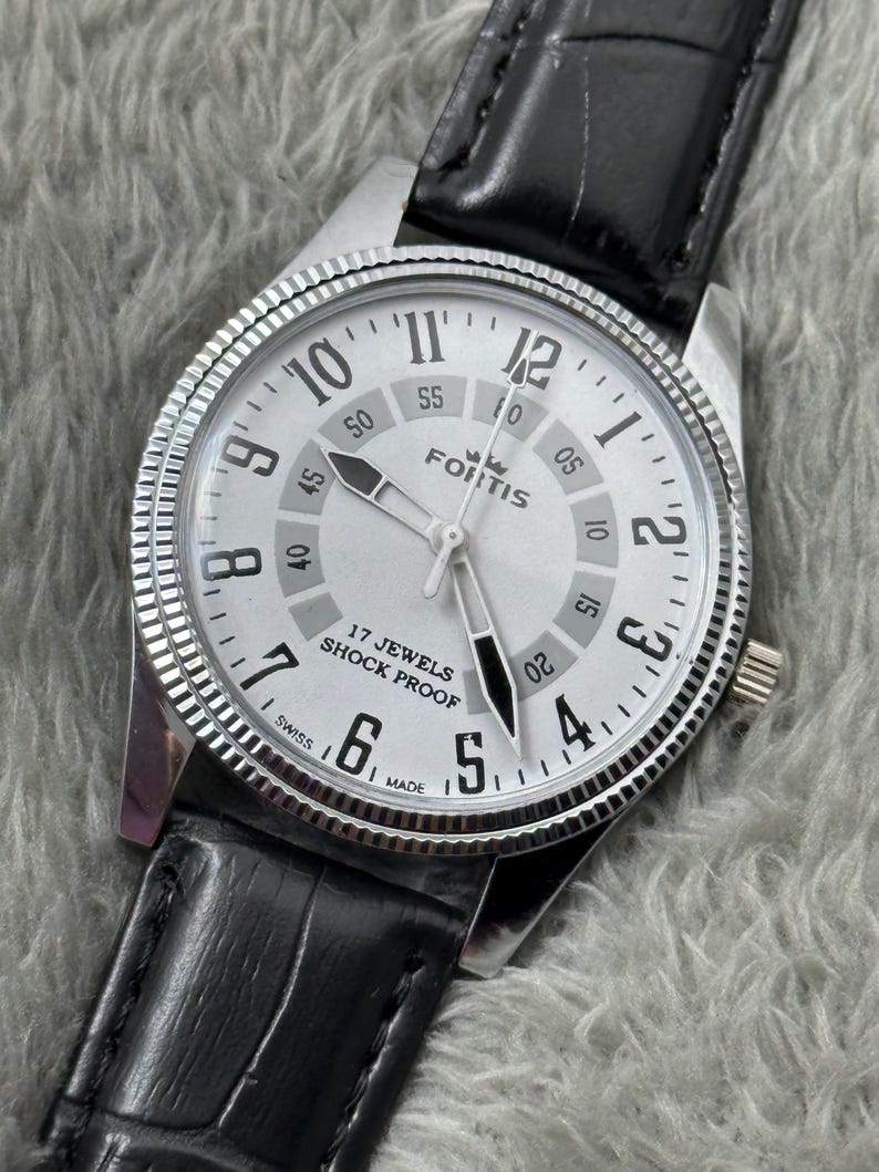 K&ouml;nnte beinhalten: Eine silberfarbene Fortis-Uhr mit wei&szlig;em Zifferblatt, schwarzen Zeigern und einem schwarzen Lederarmband. Das Zifferblatt zeigt schwarze Ziffern und den Text "17 Jewels Shock Proof". Die Uhr hat eine strukturierte L&uuml;nette.