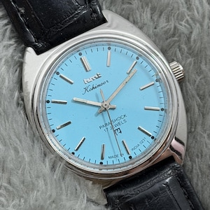 May include: A vintage Kohinoor Parashock wristwatch with a light blue face and silver hands and markers. The watch has a stainless steel case and a black leather strap. The watch face reads "Kohinoor" and "Parashock 17 Jewels".