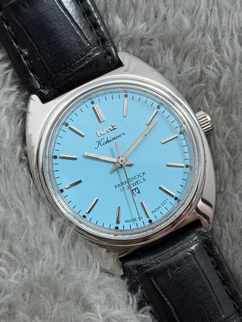 May include: A vintage Kohinoor Parashock wristwatch with a light blue face and silver hands and markers. The watch has a stainless steel case and a black leather strap. The watch face reads "Kohinoor" and "Parashock 17 Jewels".