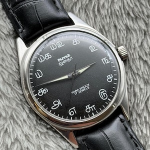 Può includere: Un orologio da polso analogico nero con cassa color argento e cinturino in pelle nera. Il quadrante dell'orologio presenta numeri bianchi e testo in lingua straniera. L'orologio indica "PARA SHOCK 17 JEWELS".