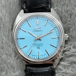 May include: A vintage HMT Kohinoor watch with a light blue face, silver case, and black leather band. The watch face features silver hands and markers, with the text "PARA SHOCK 17 JEWELS" and "MADE IN INDIA".