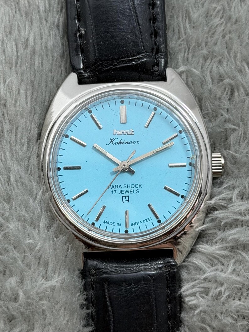 May include: A vintage HMT Kohinoor watch with a light blue face, silver case, and black leather band. The watch face features silver hands and markers, with the text "PARA SHOCK 17 JEWELS" and "MADE IN INDIA".