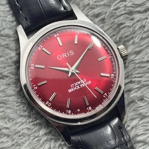 Peut inclure: Une montre-bracelet Oris vintage avec un cadran rouge vif et des aiguilles argentées. La montre a un boîtier en acier inoxydable et un bracelet en cuir noir. Le cadran affiche le nom de la marque, le texte "17 JEWELS SHOCK PROOF" et les marqueurs de temps.