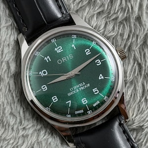 Puede incluir: Un reloj de pulsera plateado con esfera verde y correa de cuero negro. La esfera muestra la marca "ORIS", números y el texto "17 JEWELS SHOCK PROOF". El reloj tiene un diseño clásico con esfera redonda y correa de cuero texturizado.