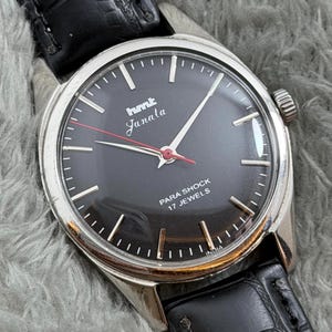 Op de afbeelding: Vintage polshorloge met een zwarte wijzerplaat, zilveren kast en zwarte leren band. De wijzerplaat toont de tekst "hmt Janala" in wit en rood, met "PARA SHOCK 17 JEWELS" eronder. Het horloge heeft een zilveren rand en een zwarte leren band.
