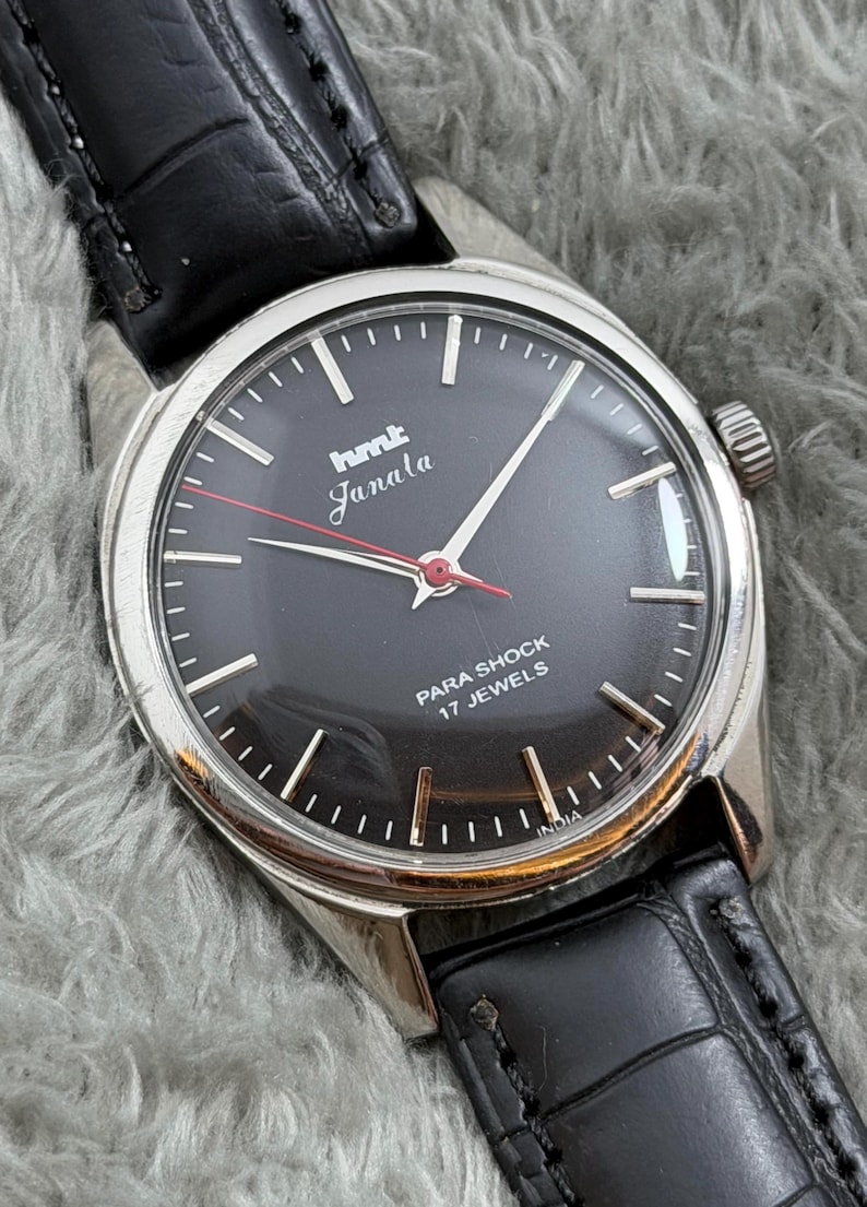 Op de afbeelding: Vintage polshorloge met een zwarte wijzerplaat, zilveren kast en zwarte leren band. De wijzerplaat toont de tekst "hmt Janala" in wit en rood, met "PARA SHOCK 17 JEWELS" eronder. Het horloge heeft een zilveren rand en een zwarte leren band.