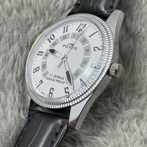 K&ouml;nnte beinhalten: Eine silberfarbene Fortis-Armbanduhr mit wei&szlig;em Zifferblatt, schwarzen Zeigern und schwarzem Lederarmband. Das Zifferblatt zeigt den Markennamen "FORTIS" und den Text "17 JEWELS SHOCK PROOF". Die Uhr hat eine strukturierte L&uuml;nette und eine Krone.