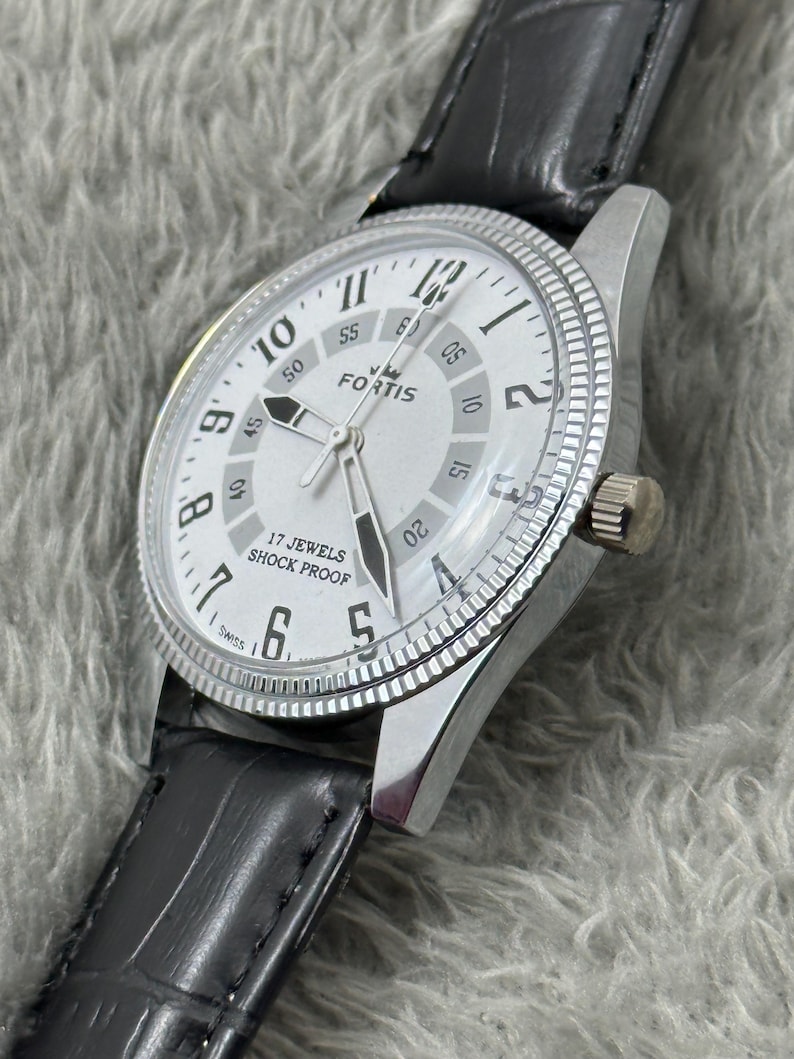 K&ouml;nnte beinhalten: Eine silberfarbene Fortis-Armbanduhr mit wei&szlig;em Zifferblatt, schwarzen Zeigern und schwarzem Lederarmband. Das Zifferblatt zeigt den Markennamen "FORTIS" und den Text "17 JEWELS SHOCK PROOF". Die Uhr hat eine strukturierte L&uuml;nette und eine Krone.