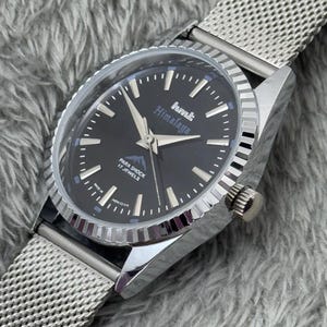 Puede incluir: Un reloj plateado con esfera negra y correa de malla. La esfera del reloj muestra el texto "hmt Himalaya" y "Para Shock 17 Jewels". El reloj tiene agujas y marcadores plateados.