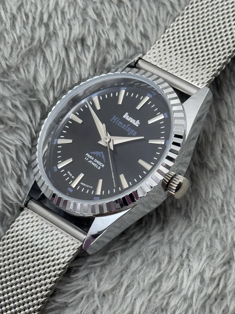Puede incluir: Un reloj plateado con esfera negra y correa de malla. La esfera del reloj muestra el texto "hmt Himalaya" y "Para Shock 17 Jewels". El reloj tiene agujas y marcadores plateados.
