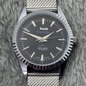 Puede incluir: Un reloj de pulsera plateado con esfera negra y correa de malla. La esfera del reloj muestra el texto "hmt Himalaya" y "PARA SHOCK 17 JEWELS". El reloj tiene agujas e indicadores plateados.