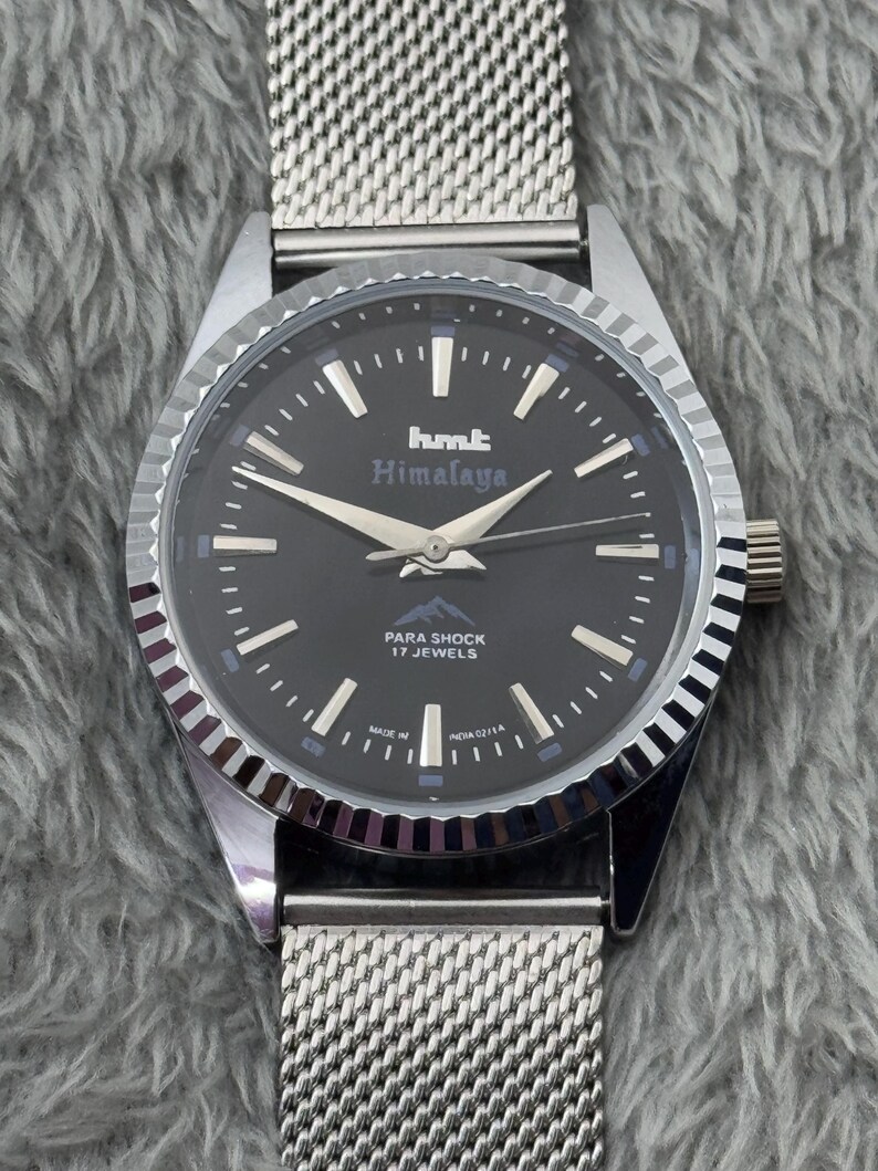 Puede incluir: Un reloj de pulsera plateado con esfera negra y correa de malla. La esfera del reloj muestra el texto "hmt Himalaya" y "PARA SHOCK 17 JEWELS". El reloj tiene agujas e indicadores plateados.