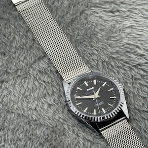 Puede incluir: Un reloj de pulsera con esfera negra y agujas e indicadores plateados. El reloj tiene una correa de malla plateada y un bisel plateado. La esfera del reloj muestra el texto "Himalaya" y "21 JEWELS".
