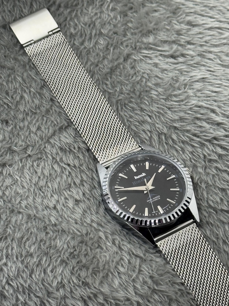 Puede incluir: Un reloj de pulsera con esfera negra y agujas e indicadores plateados. El reloj tiene una correa de malla plateada y un bisel plateado. La esfera del reloj muestra el texto "Himalaya" y "21 JEWELS".