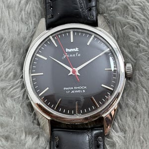 Op de afbeelding: Een vintage polshorloge met een zwarte wijzerplaat, zilveren kast en zwarte leren band. De wijzerplaat heeft de tekst "hmt Janata" en "PARA SHOCK 17 JEWELS". Het horloge heeft zilveren wijzers en een rode secondewijzer.