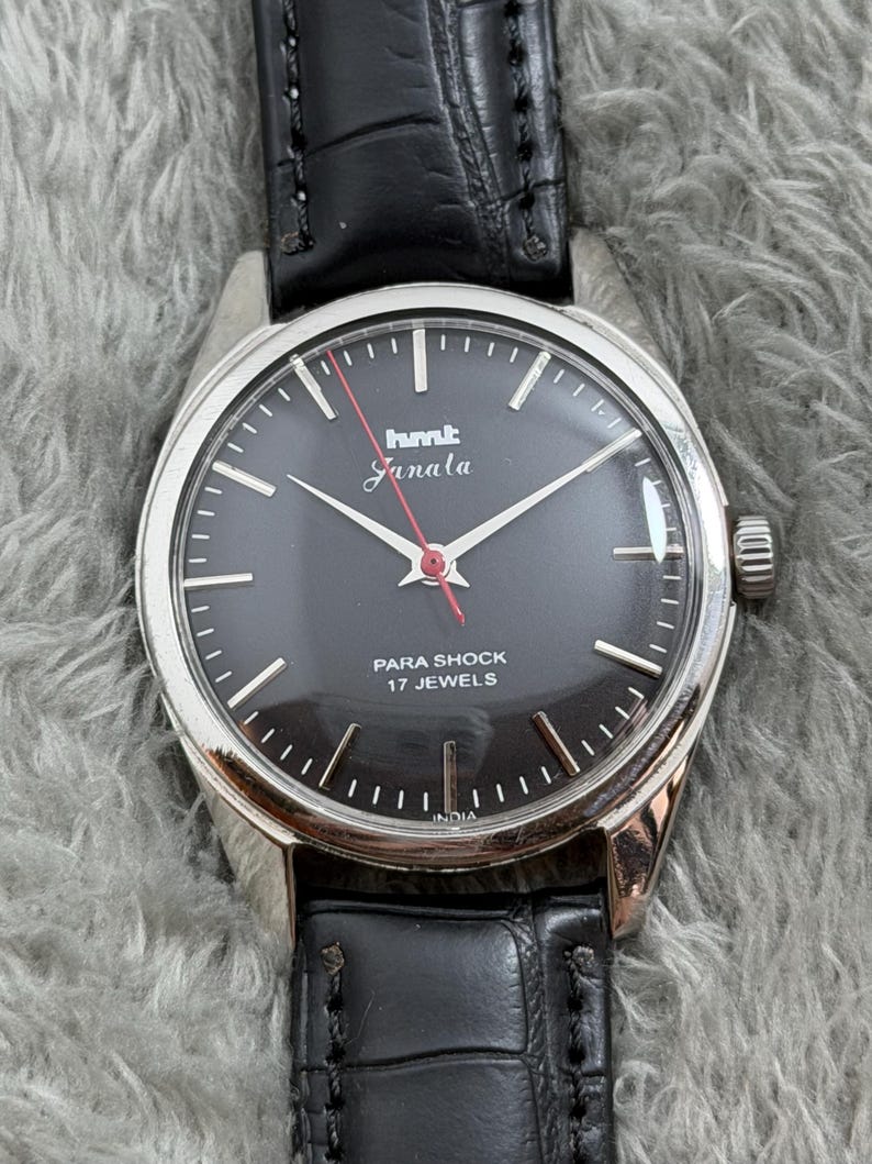 Op de afbeelding: Een vintage polshorloge met een zwarte wijzerplaat, zilveren kast en zwarte leren band. De wijzerplaat heeft de tekst "hmt Janata" en "PARA SHOCK 17 JEWELS". Het horloge heeft zilveren wijzers en een rode secondewijzer.
