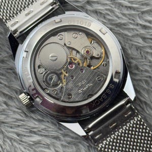 Puede incluir: Primer plano de un reloj de pulsera plateado con una correa de malla, que muestra los intrincados mecanismos internos. La parte trasera del reloj muestra engranajes, joyas y el texto "S.S. BACK" y "011019". El reloj est&aacute; sobre un fondo gris texturizado.