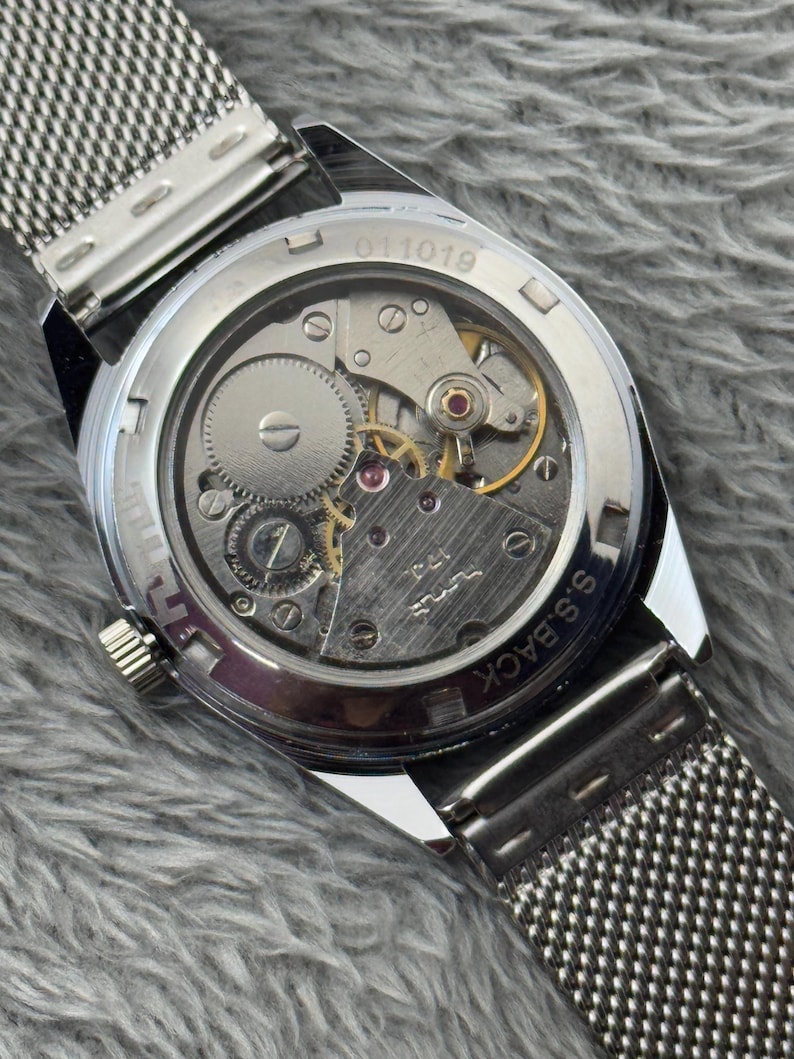 Puede incluir: Primer plano de un reloj de pulsera plateado con una correa de malla, que muestra los intrincados mecanismos internos. La parte trasera del reloj muestra engranajes, joyas y el texto "S.S. BACK" y "011019". El reloj est&aacute; sobre un fondo gris texturizado.