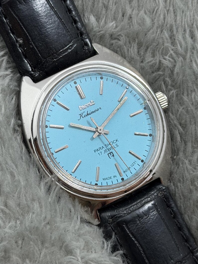 May include: A vintage Kohinoor Para Shock watch with a light blue face and silver hands and markers. The watch has a stainless steel case and a black leather strap. The watch face includes the text "PARA SHOCK 17 JEWELS" and "MADE IN INDIA 0231".