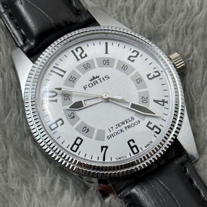 K&ouml;nnte beinhalten: Eine silberfarbene Fortis-Armbanduhr mit wei&szlig;em Zifferblatt, schwarzen Zeigern und einem schwarzen Lederarmband. Das Zifferblatt zeigt den Markennamen, "17 Jewels Shock Proof" und "Swiss". Die Uhr hat eine strukturierte L&uuml;nette.