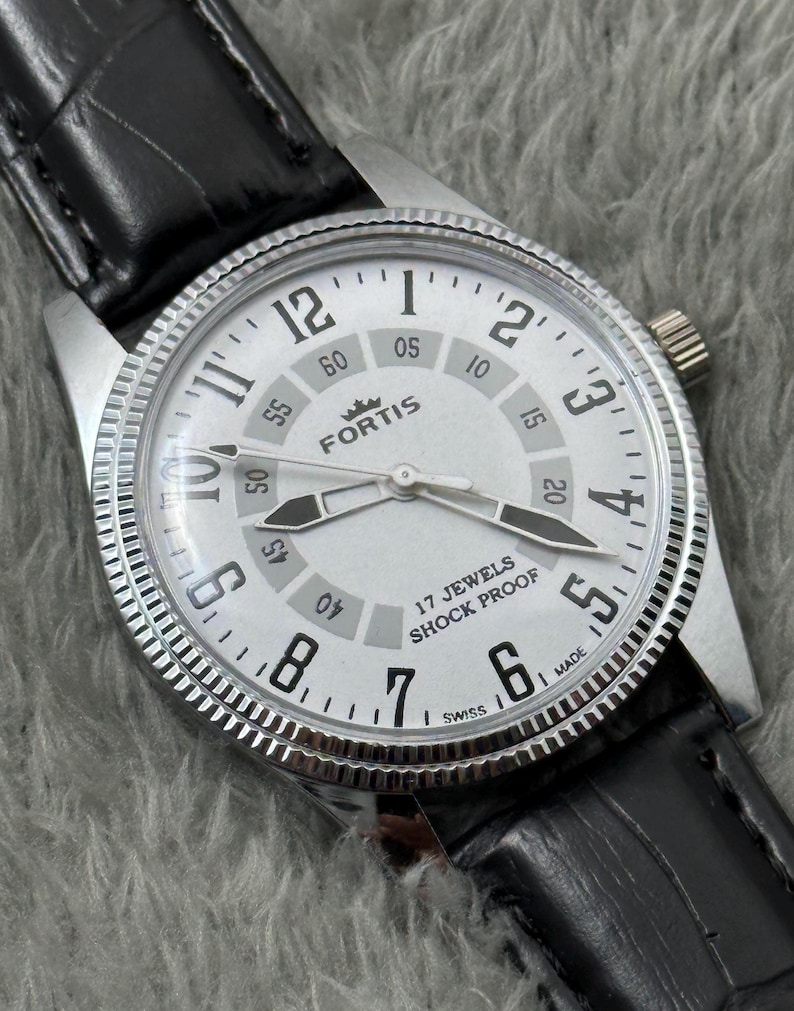 K&ouml;nnte beinhalten: Eine silberfarbene Fortis-Armbanduhr mit wei&szlig;em Zifferblatt, schwarzen Zeigern und einem schwarzen Lederarmband. Das Zifferblatt zeigt den Markennamen, "17 Jewels Shock Proof" und "Swiss". Die Uhr hat eine strukturierte L&uuml;nette.