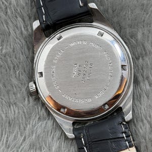 Op de afbeelding: Close-up van een vintage polshorloge, met de roestvrijstalen achterkant met tekst die water- en stofbescherming aangeeft. Het horloge heeft een zwarte leren band met witte stiksels. Het horloge is schokbestendig.