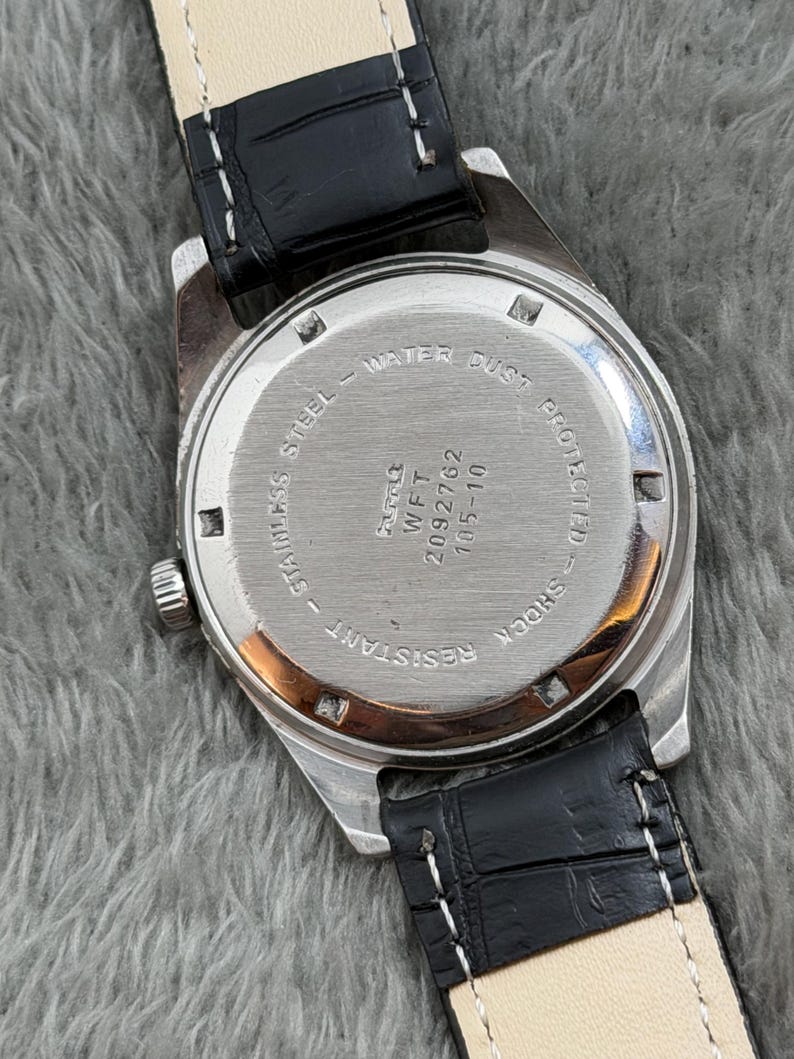 Op de afbeelding: Close-up van een vintage polshorloge, met de roestvrijstalen achterkant met tekst die water- en stofbescherming aangeeft. Het horloge heeft een zwarte leren band met witte stiksels. Het horloge is schokbestendig.