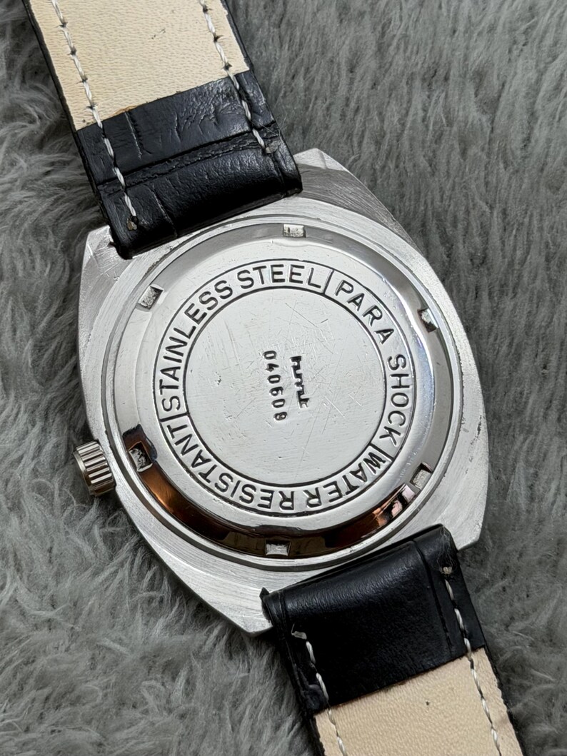 May include: A stainless steel watch with a black leather band. The watch face is silver with the words "Stainless Steel", "Water Resistant", and "Para Shock" engraved. The watch band is black leather with white stitching.