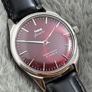 Peut inclure: Gros plan d'une montre-bracelet vintage avec un cadran bordeaux et un boîtier argenté. La montre porte l'inscription "hmt Janala" et "PARA SHOCK 17 JEWELS". Elle est dotée d'un bracelet en cuir noir.