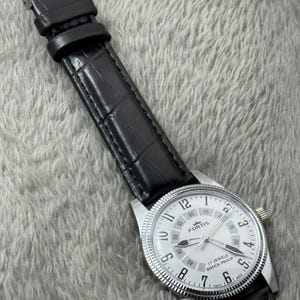 K&ouml;nnte beinhalten: Eine silberfarbene Fortis-Uhr mit wei&szlig;em Zifferblatt, schwarzen Zeigern und einem schwarzen Lederarmband. Das Zifferblatt zeigt die Zahl 12 und den Text "17 JEWELS SHOCK PROOF". Das Armband hat ein Krokodilmuster.