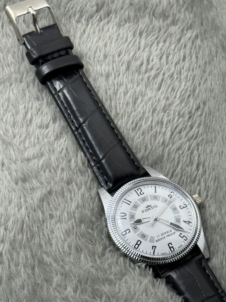 K&ouml;nnte beinhalten: Eine silberfarbene Fortis-Uhr mit wei&szlig;em Zifferblatt, schwarzen Zeigern und einem schwarzen Lederarmband. Das Zifferblatt zeigt die Zahl 12 und den Text "17 JEWELS SHOCK PROOF". Das Armband hat ein Krokodilmuster.