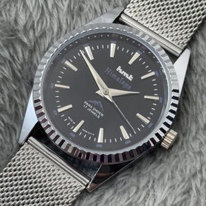 Puede incluir: Un reloj de pulsera plateado con esfera negra y correa de malla. La esfera del reloj muestra el texto "hmt Himalaya" y "PARA SHOCK 17 JEWELS". El reloj tiene agujas e indicadores plateados.