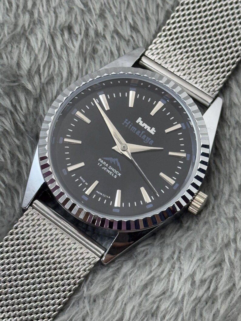 Puede incluir: Un reloj de pulsera plateado con esfera negra y correa de malla. La esfera del reloj muestra el texto "hmt Himalaya" y "PARA SHOCK 17 JEWELS". El reloj tiene agujas e indicadores plateados.