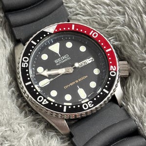 Peut inclure: Une montre de plongée automatique Seiko noire avec un cadran noir, des marqueurs blancs et une lunette rotative rouge et noire. La montre a un bracelet en caoutchouc noir et un boîtier en acier inoxydable.