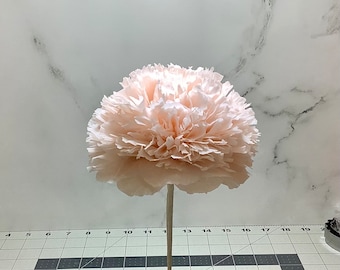 May include: A large, light peach-colored paper flower with ruffled petals. The flower is mounted on a thin, light-colored stem, and is set in a small, round, light brown base. The background is a white marble pattern.