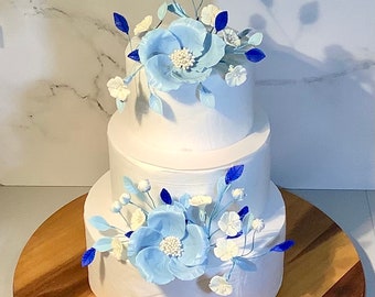 May include: A three-tiered white cake decorated with light blue and white floral accents. The cake sits on a wooden cake stand. The flowers have a detailed design, with blue petals and white centers.