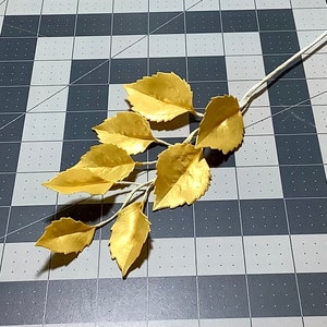 May include: A sprig of gold-painted leaves on a white stem. The leaves are arranged in a cascading pattern, with the largest leaf at the top and the smallest leaves at the bottom.