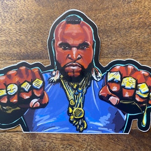 May include: A sticker featuring a cartoon illustration of Mr. T, a popular 1980s television character. He is wearing a blue shirt, gold chains, and multiple gold rings on his fingers. He is looking directly at the viewer with a serious expression.