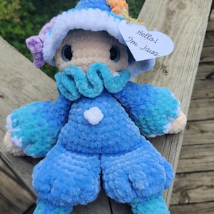May include: A plush crochet doll featuring a blue and white color scheme with a ruffled collar, a pointed hat, and a white heart-shaped tag attached that reads Hello! Im Java. The doll has large dark eyes and a soft, textured yarn construction.