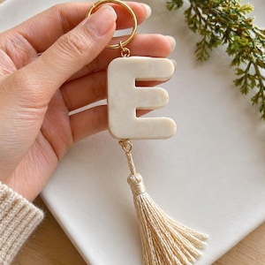 May include: Cream-colored letter "E" keychain with a gold-tone keyring and tassel. The letter is a solid, blocky design. The tassel is made of cream-colored threads. The keychain is held by a hand.