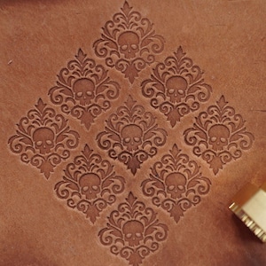 May include: A close-up of a leather craft project with a repeating skull and scroll pattern stamped into brown leather. A metal stamp tool with a brass head is in the lower right corner.