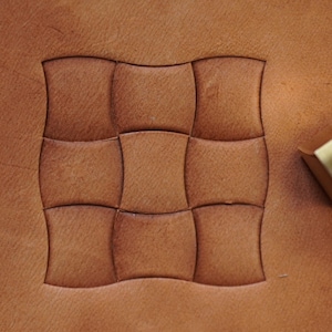 May include: A close-up of brown leather showing a stamped basketweave design. A metal and brass leather stamping tool is positioned nearby, indicating leather crafting. The leather has a natural, textured appearance, with the pattern clearly defined.