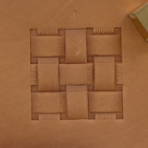 May include: Close-up of a tan leather surface with a woven basket pattern embossed in the center. A brass tool is in the upper right corner, likely used for the embossing process. The leather has a textured appearance.