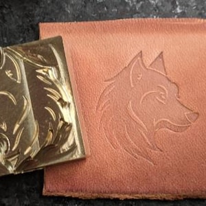 May include: A square, gold-colored metal stamp with a wolf head design next to a piece of brown leather with the same wolf head embossed on it. The stamp is square and the leather is rectangular.
