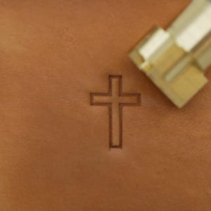 May include: A close-up of a brown leather surface with a cross imprint. A brass tool is positioned at the top right corner. The cross is a simple, outlined design, and the leather has a smooth texture.