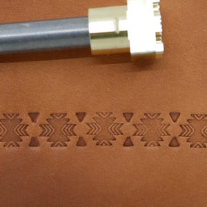 Op de afbeelding: Een leermarkeergereedschap met een geometrisch ontwerp wordt getoond naast een gestempeld patroon op bruin leer. De tool heeft een metalen handvat en een messing stempelkop. Het gestempelde patroon heeft herhaalde geometrische vormen.
